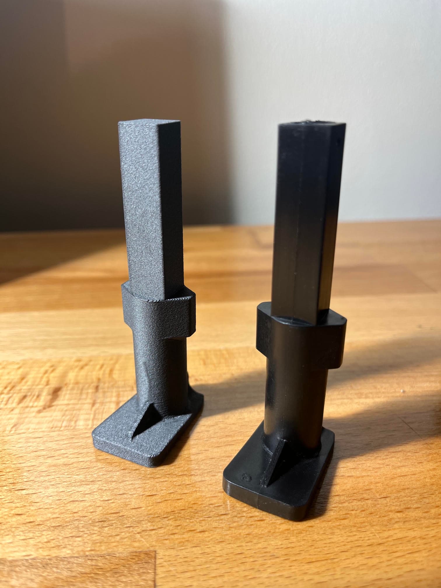 Deux pièces en plastique noir identiques, l'une mate et l'autre brillante, sur une surface boisée.