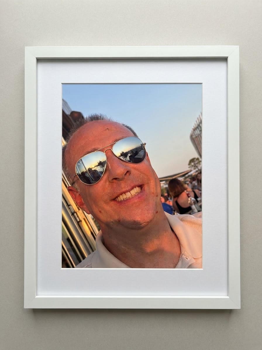 Portrait encadré d'un homme souriant avec des lunettes aviateur reflétant un coucher de soleil.
